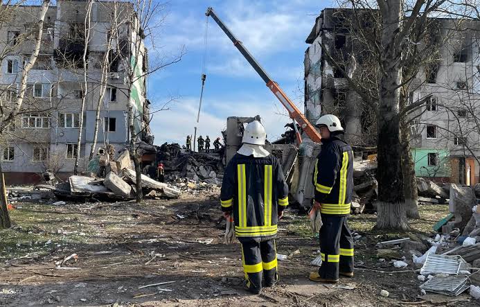 Рятувальники ДСНС у захисних касках і форменому одязі на тлі зруйнованого внаслідок ворожого обстрілу житлового будинку та будівельної техніки.